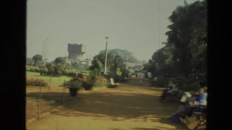1974 India park view with a dirt path, manicured planting, benches, trees, and visitors sitting or walking nearby.