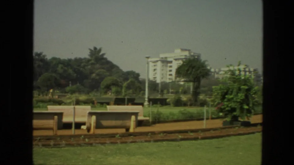 1974 India 8mm frame showing benches along a landscaped park, with trees and apartment buildings visible beyond the path.