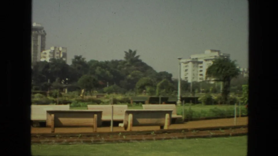 1974 8mm India park view with benches, planted beds, trees, lawns, and modern buildings in the distance.