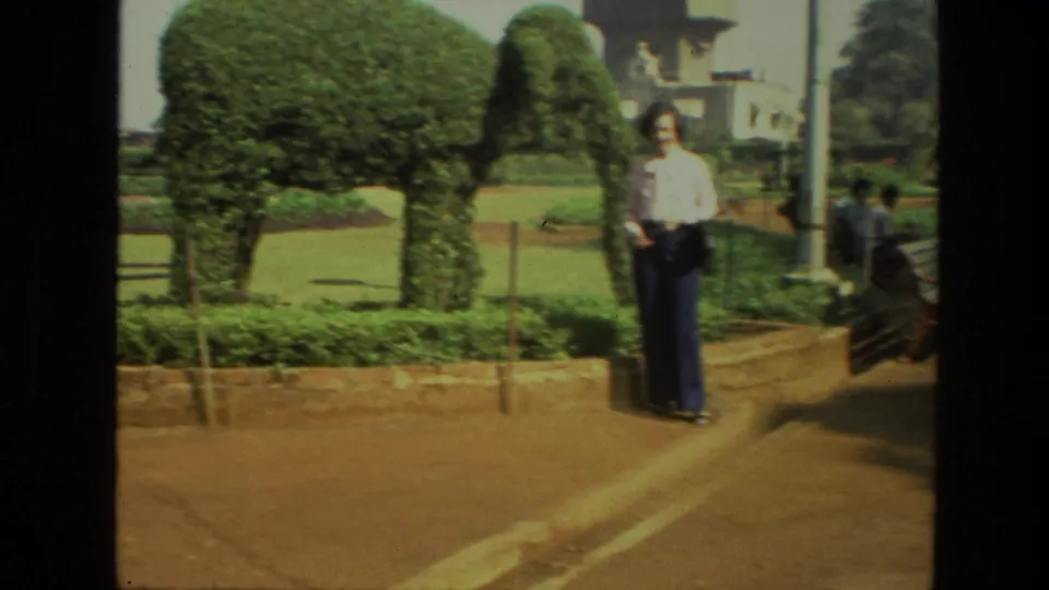A man stands beside elephant-shaped topiary in a 1974 India park, with paths, lawns, and other visitors nearby.