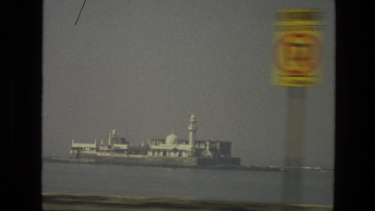 Distant island or waterfront building complex in India, seen across water with a blurred roadside sign in the foreground.