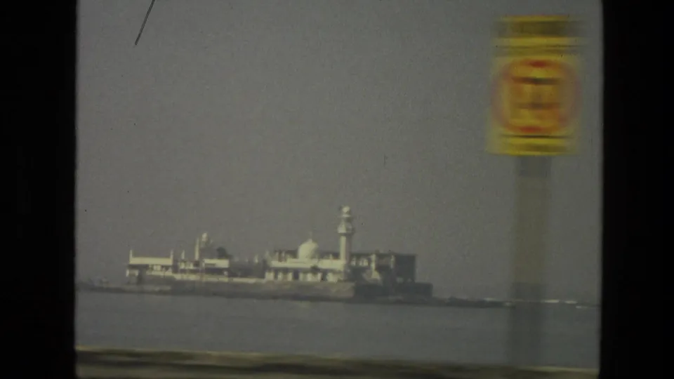 Distant island or waterfront building complex in India, seen across water with a blurred roadside sign in the foreground.