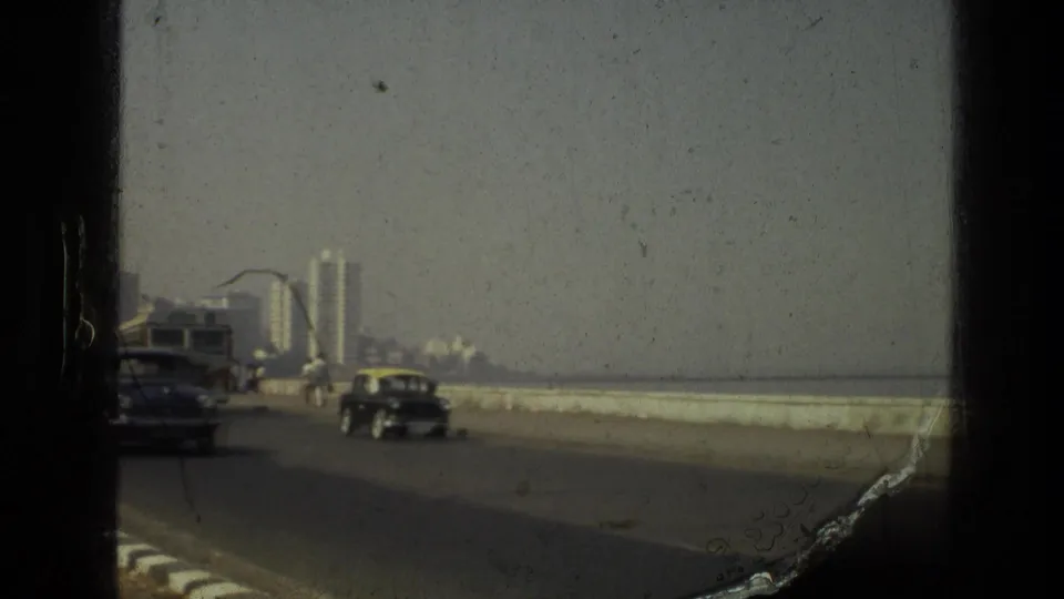 1974 India home-movie frame of cars traveling along a waterfront road with a sea wall and distant buildings ahead.