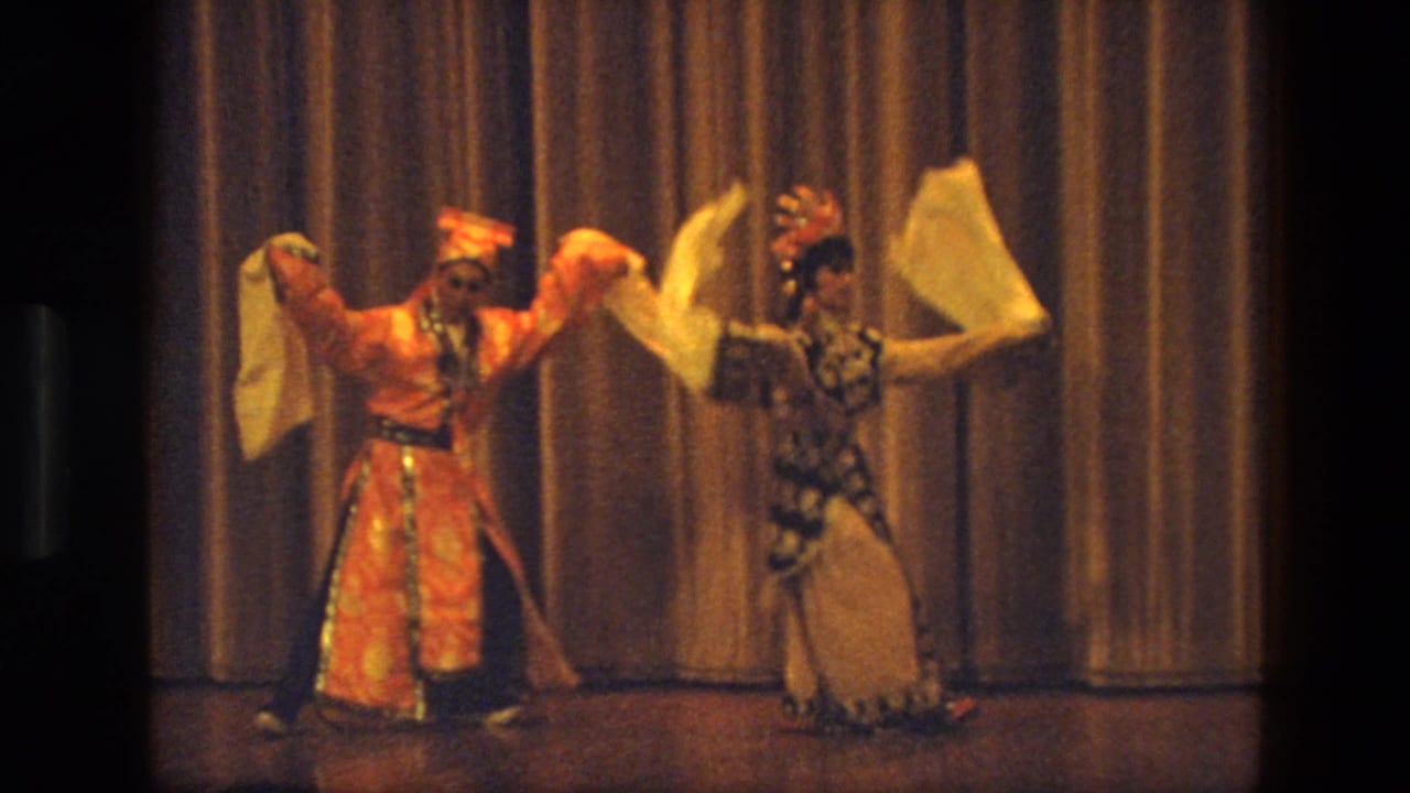 Two costumed adult performers dance on a stage with white scarves before a gold curtain.
