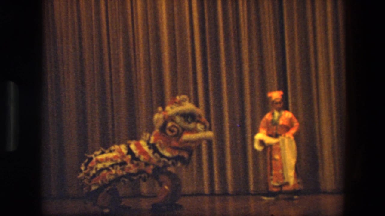 Lion dance costume and costumed performer on a stage before a gold curtain.