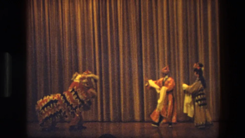 1974 India 8mm frame with a lion costume at left and two costumed performers crossing the stage before a curtain.