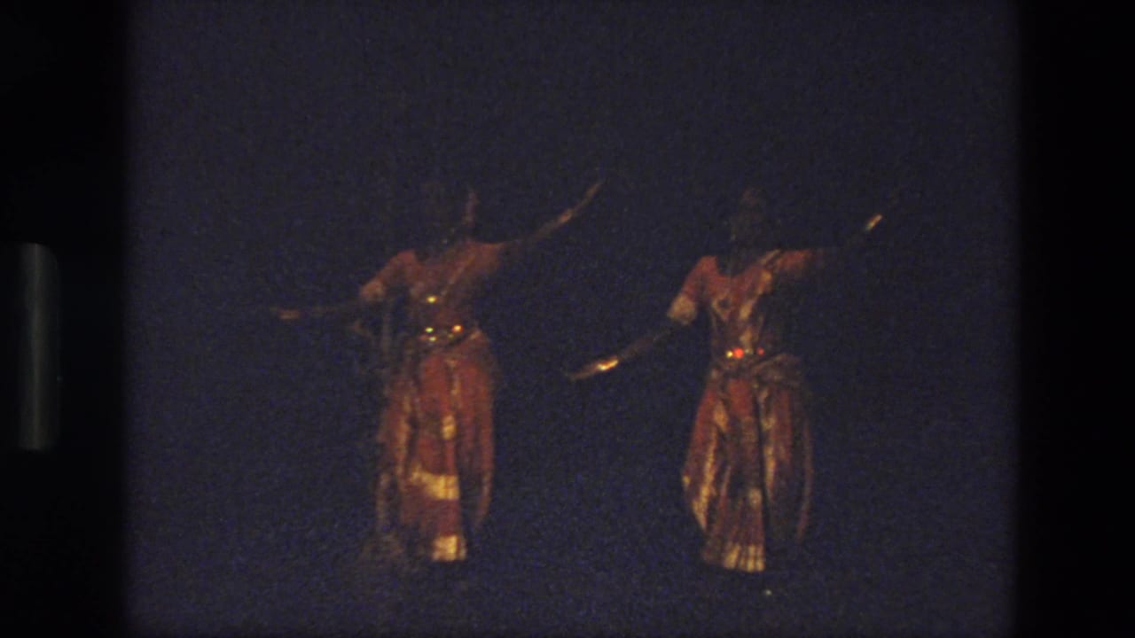 Two women perform on a dark stage in India, 1974, wearing red-and-gold costumes with reflective trim and extended arm poses.