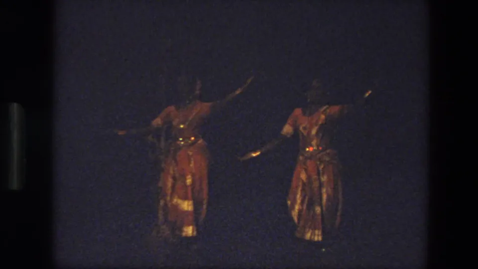 Two women perform on a dark stage in India, 1974, wearing red-and-gold costumes with reflective trim and extended arm poses.
