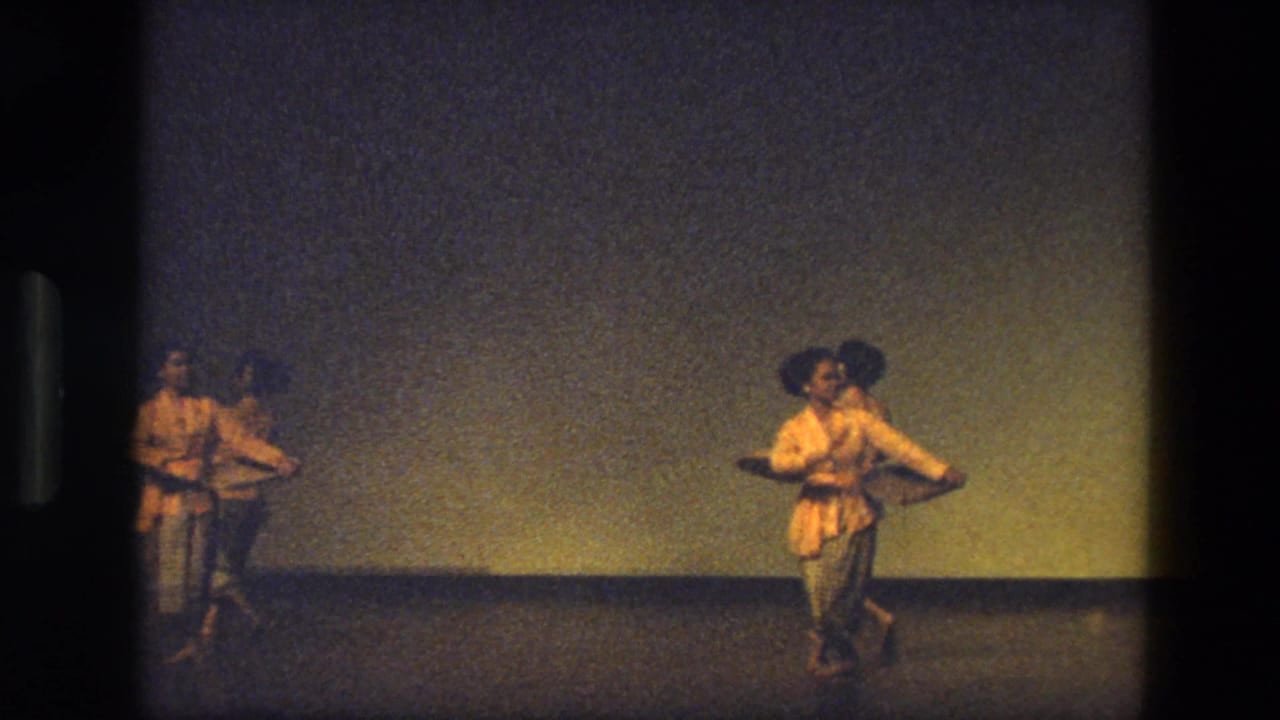1974 India 8mm frame showing dancers in light traditional-looking costumes spread across a dim stage with a plain backdrop.