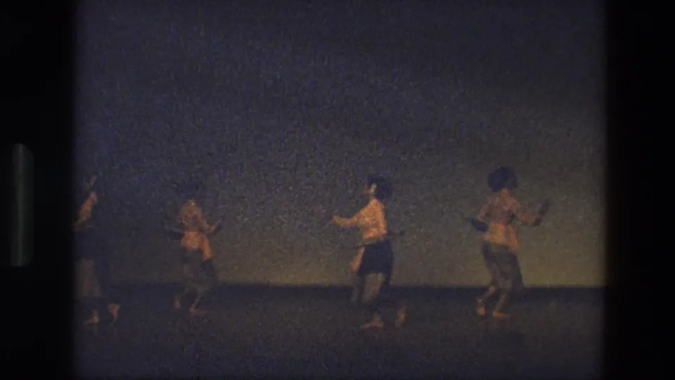 1974 India 8mm frame showing three dancers in motion on a dark stage, captured from an audience perspective with heavy grain.