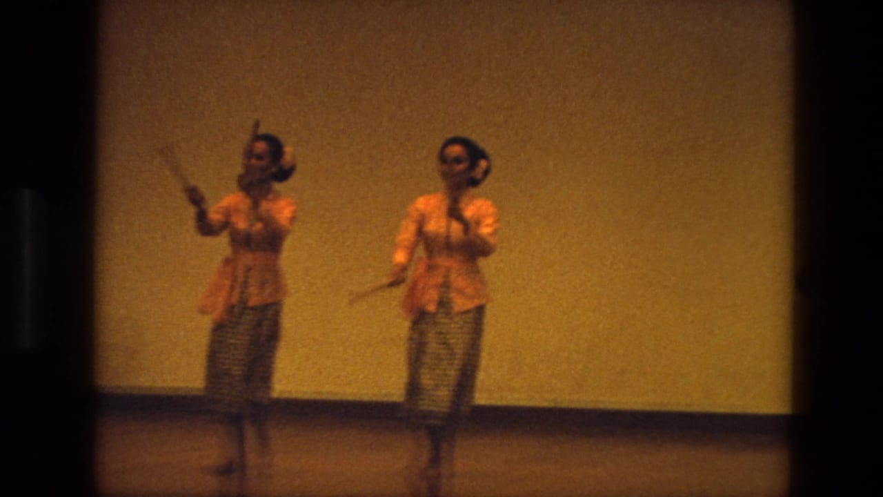 In 1974 India footage, two women in orange blouses and patterned skirts perform with wooden clappers on a dim indoor stage.