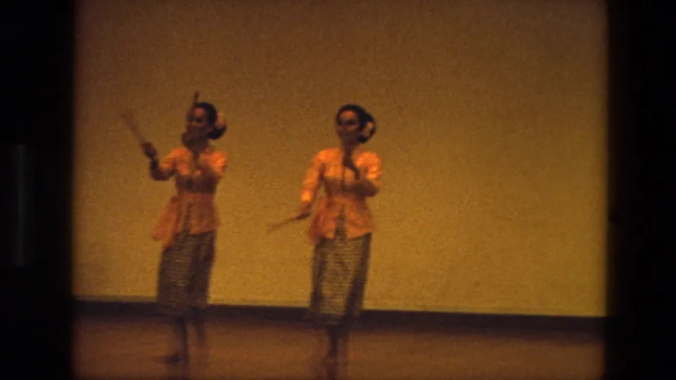 In 1974 India footage, two women in orange blouses and patterned skirts perform with wooden clappers on a dim indoor stage.
