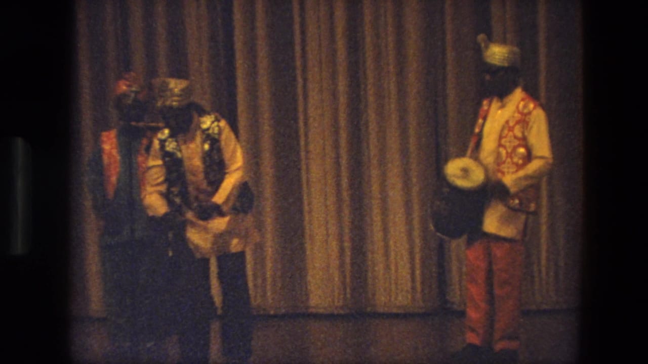 Costumed male performers stand near a curtain with a drummer at right.