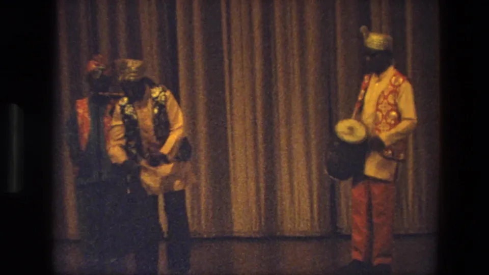 Costumed male performers on an indoor stage, with one drummer at right and others grouped near a curtain in 1974 India footage.