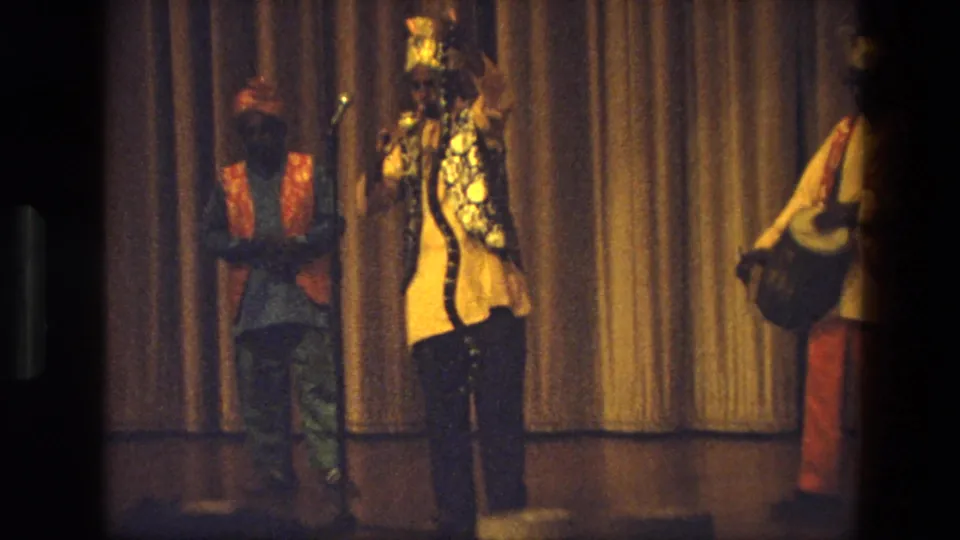 A central performer in ornate costume and headwear appears to sing into a microphone, flanked by musicians on a curtained stage in India.