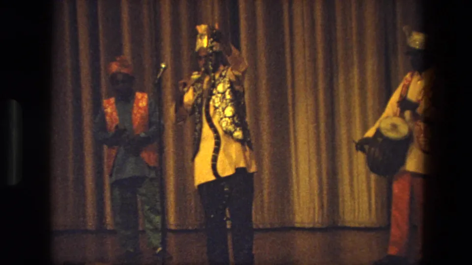 The ensemble remains on a curtained stage, with a central singer at the microphone and a drummer partly visible at right.