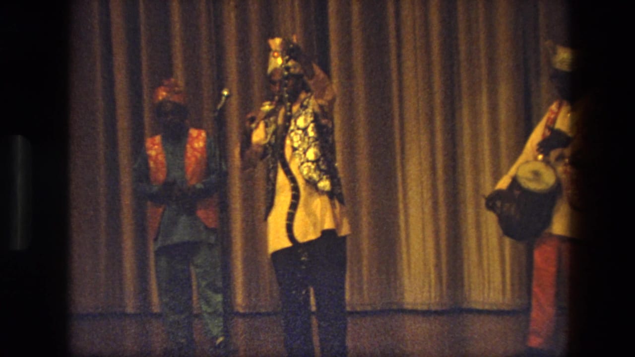 1974 India home-movie frame of three performers on a dim stage, including a singer at a microphone, a musician, and a drummer.