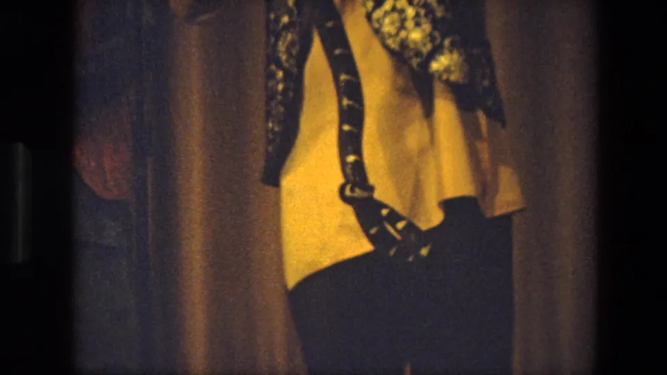 1974 India home-movie frame with a tight crop of a costumed performer standing before warm stage curtains.