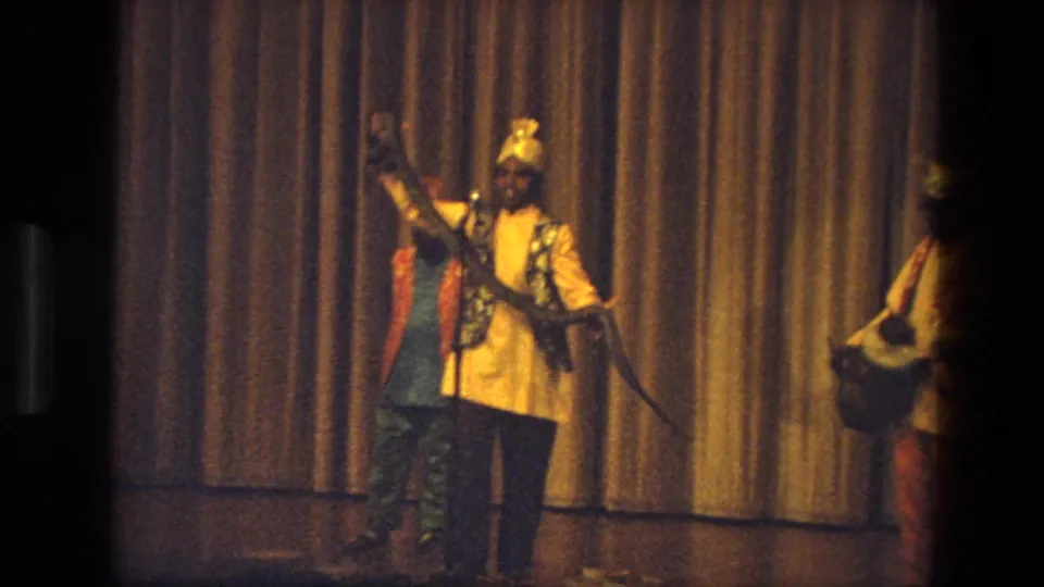 1974 India 8mm frame showing a man in traditional-style attire holding a snake on stage, with another performer partly visible at right.