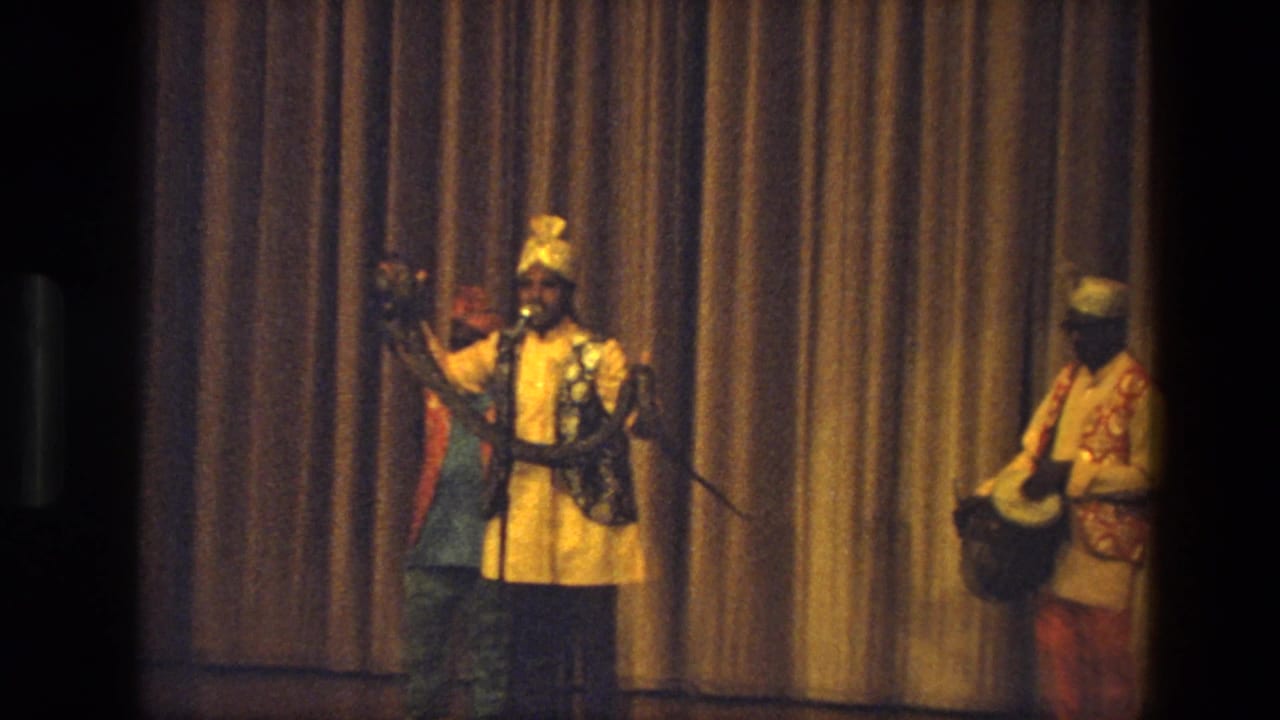Wide stage view of a man in ornate attire holding a snake near a microphone while a drummer stands nearby.