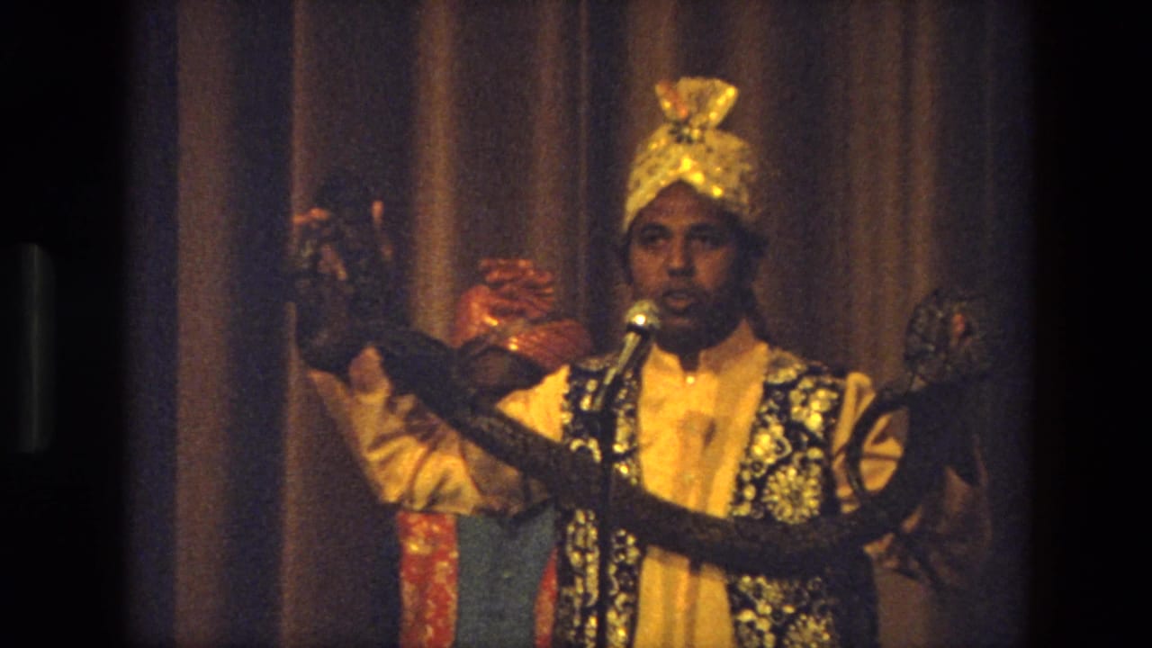 Close 1974 India stage view of a costumed performer addressing the audience with a microphone and displaying a snake.