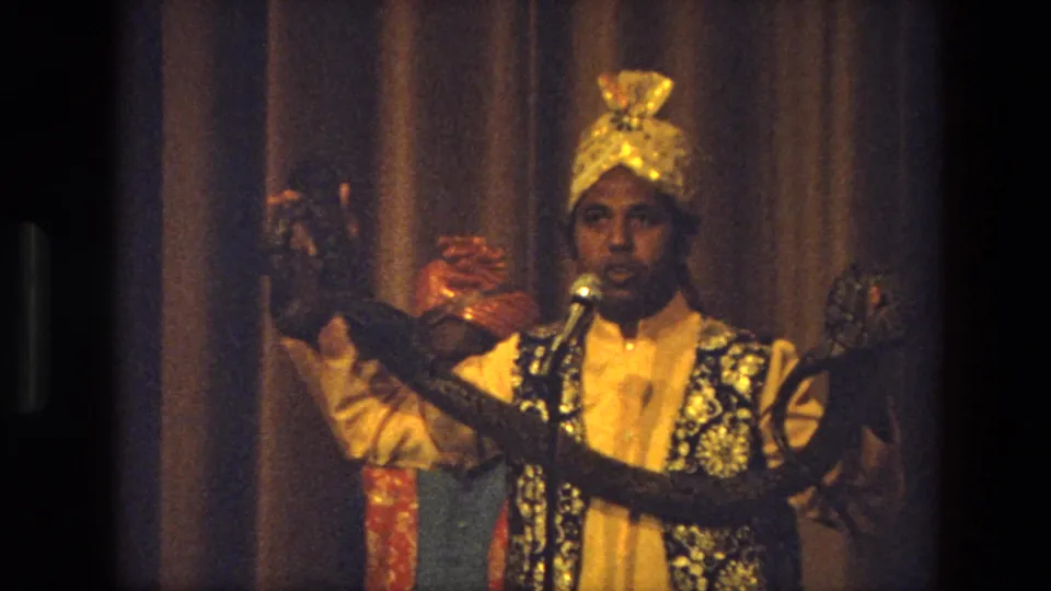 Close 1974 India stage view of a costumed performer addressing the audience with a microphone and displaying a snake.