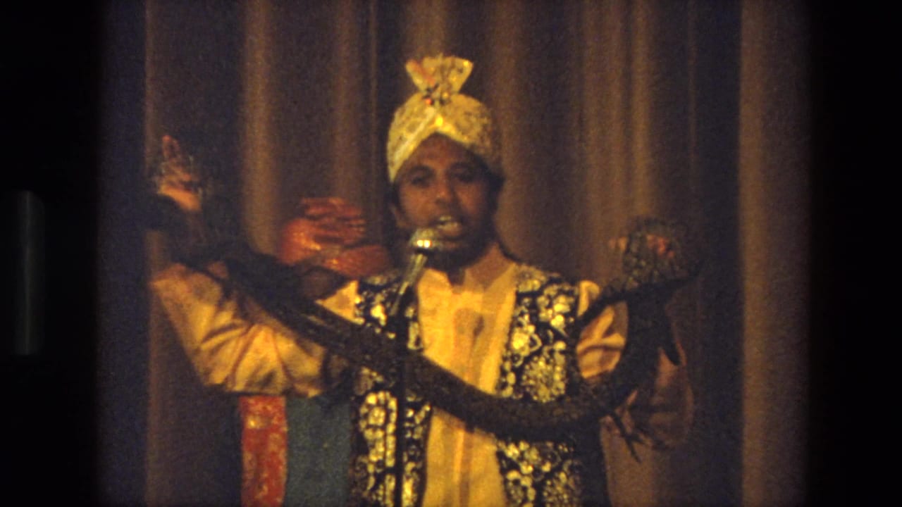 A 1974 India stage performer in a gold turban holds a long snake while speaking or singing into a microphone.