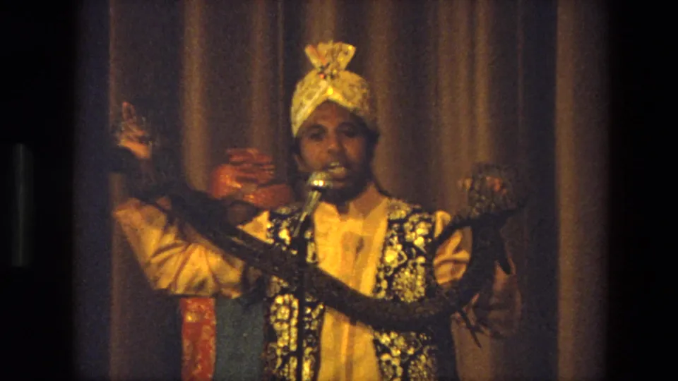 A 1974 India stage performer in a gold turban holds a long snake while speaking or singing into a microphone.