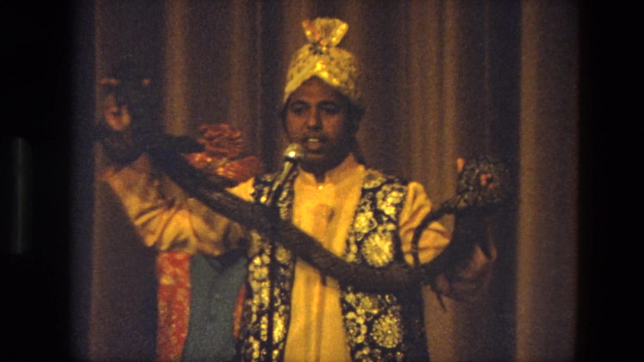 Final close view of the 1974 India stage act, with the performer holding the snake across his costume under warm lights.