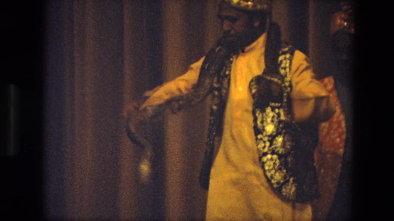 1974 India 8mm frame of a performer in a golden turban and patterned vest guiding a snake across his shoulder.