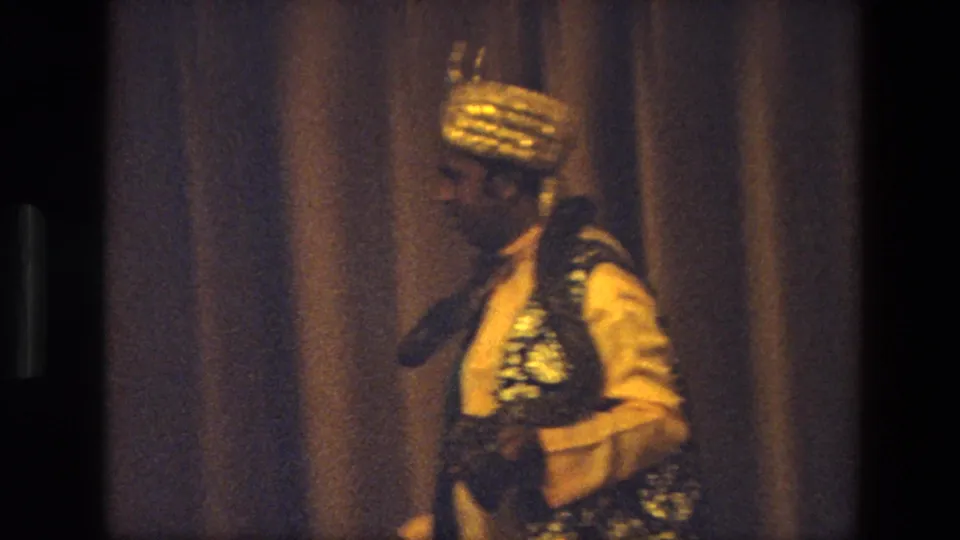 1974 India 8mm frame of the performer in profile, wearing golden headwear and an ornate vest while handling the snake.