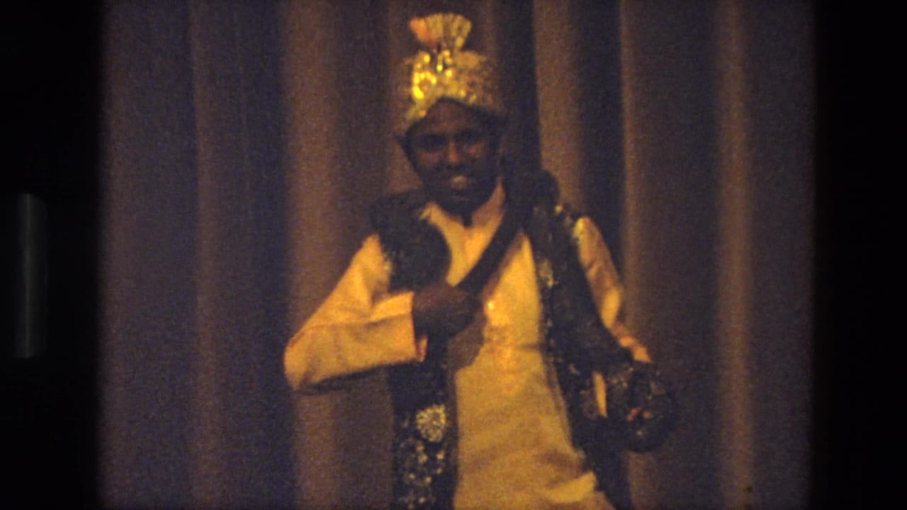 Snake handler in ornate costume stands on a stage holding a coiled snake.
