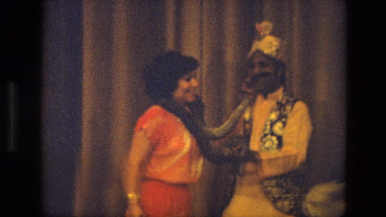 Woman in a red sari stands beside a snake handler on stage.