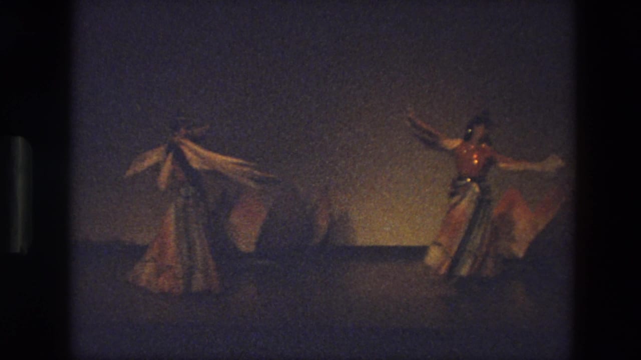 Three dancers on a dim stage, two holding long scarves while one turns in a full dress.