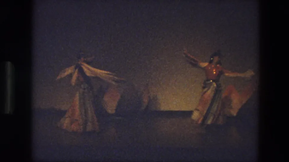 1974 India 8mm frame showing three stage dancers in flowing dresses, with long scarves raised under warm low light.