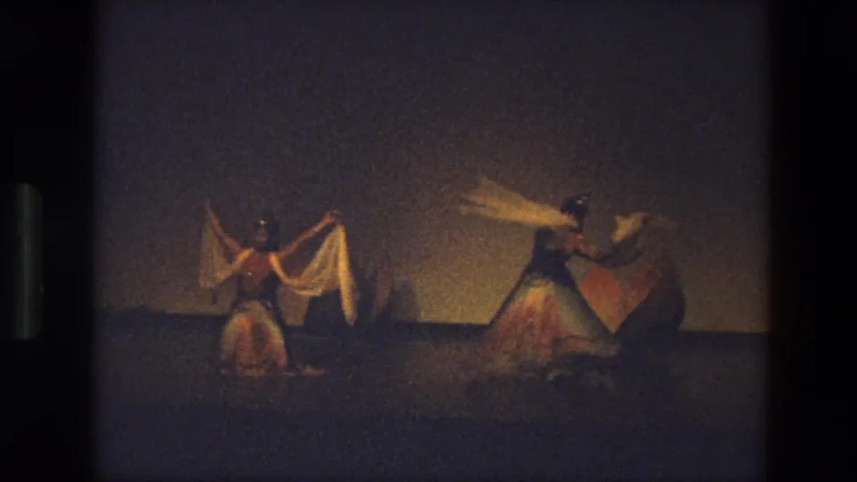 1974 India 8mm frame of three dancers spaced across a low-lit stage, holding scarves outward during a synchronized turn.