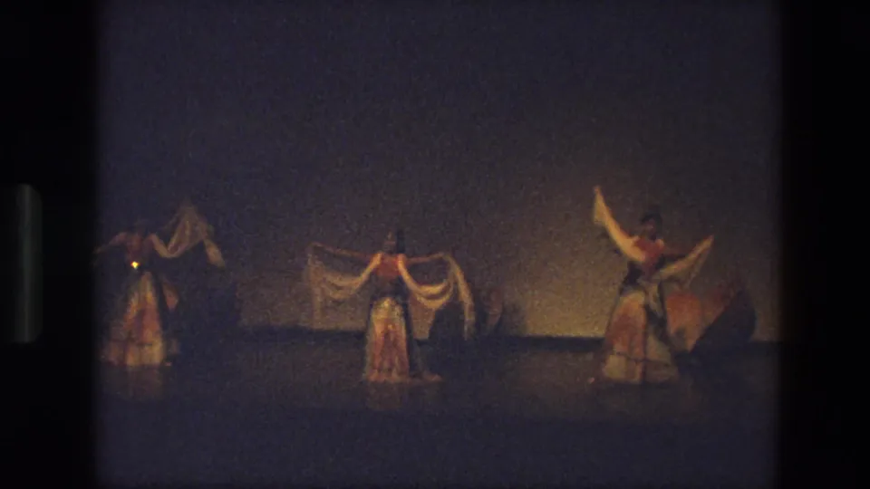 1974 India 8mm frame showing a low-light stage dance with three visible performers, flowing skirts, and pale scarves.