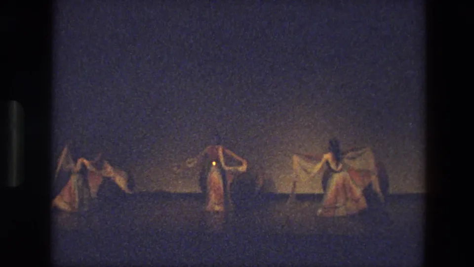 1974 India 8mm frame with three stage dancers holding scarves in a wide formation under subdued warm lighting.