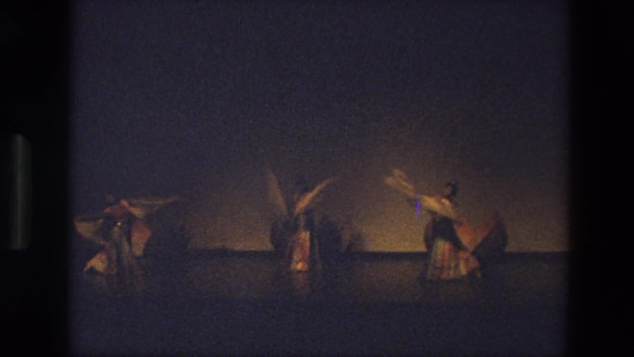 In 1974 India footage, three dancers in warm-toned costumes perform on a dim stage with veils raised and spread.