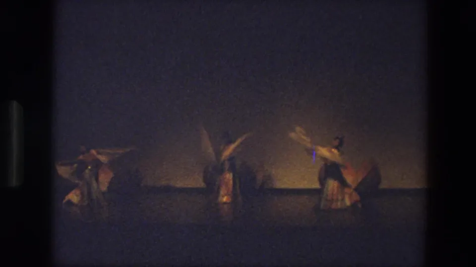 In 1974 India footage, three dancers in warm-toned costumes perform on a dim stage with veils raised and spread.