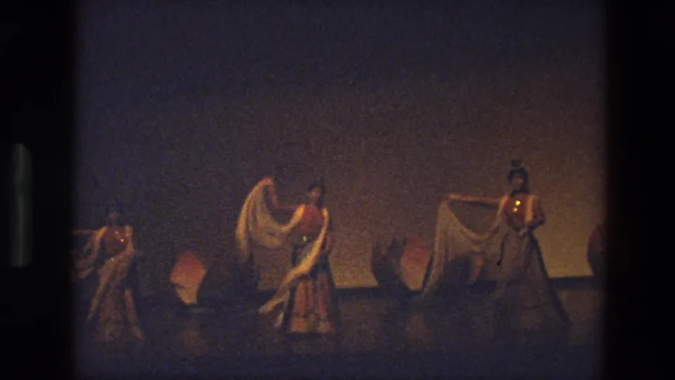 1974 India 8mm frame showing three women performing a staged dance with white scarves before pink lotus flower props.