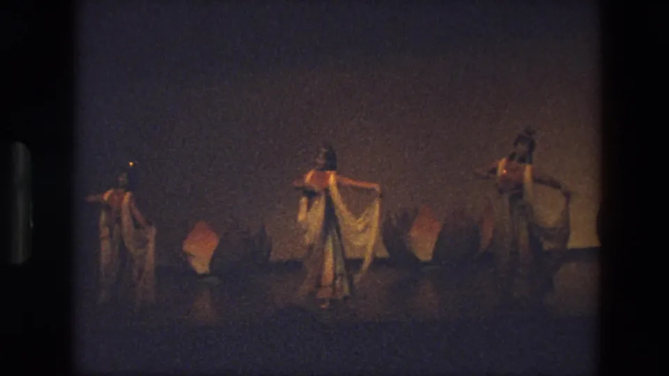 Three women appear spread across a low-lit stage, continuing a scarf dance among lotus-shaped props in 1974 India footage.