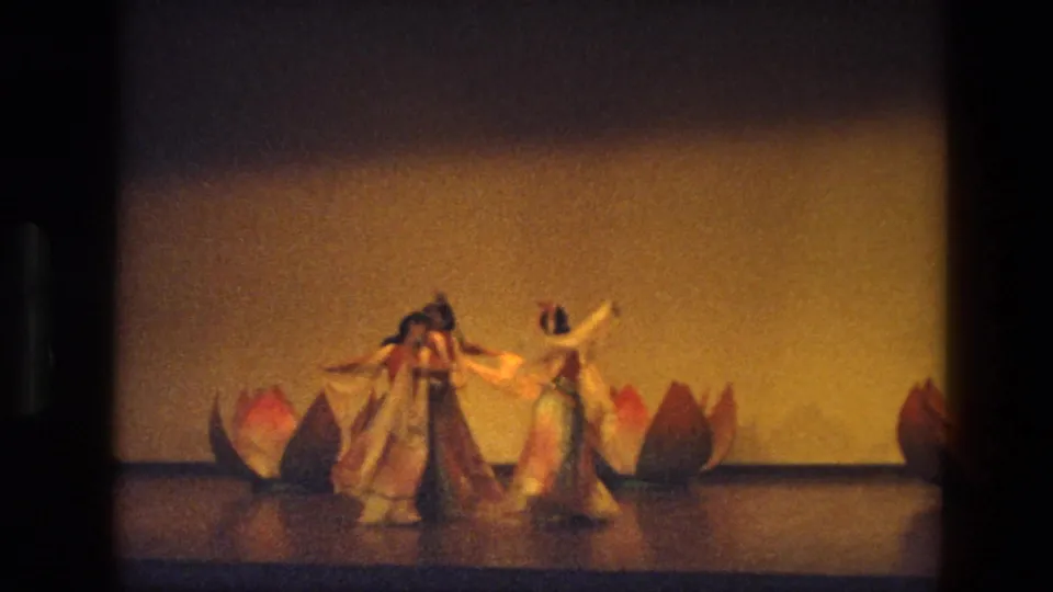 A warmer, brighter frame from the 1974 India dance clip shows two women centered with scarves and lotus props behind them.