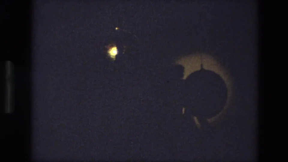 1974 India 8mm frame showing a dim stage with a crescent-like shadow form and a small warm light above it.