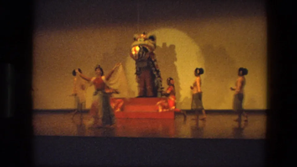 1974 India home-movie frame showing a dancer with raised arms beside a red platform and large lion-like stage costume.