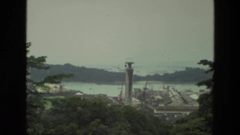 1974 India 8mm frame showing a hazy waterway, wooded foreground, distant buildings, and a tall pillar-like structure near the shore.