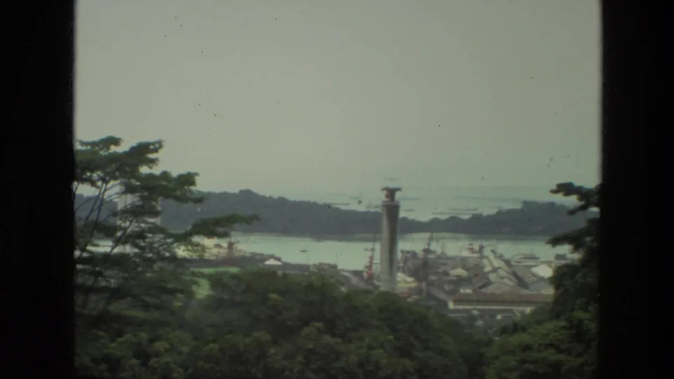 1974 India home-movie frame with trees partly obscuring a reservoir or dam area, a tall vertical structure, and distant water under haze.