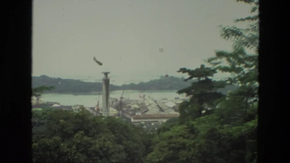 A 1974 India home-movie frame reveals a coastal town below, with trees in front, a tall tower, water, and hazy hills in the distance.