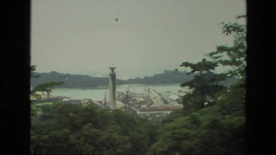 1974 India home-movie frame looking from a hillside through dark edges toward trees, a harbor, rooftops, water, and a tall tower.