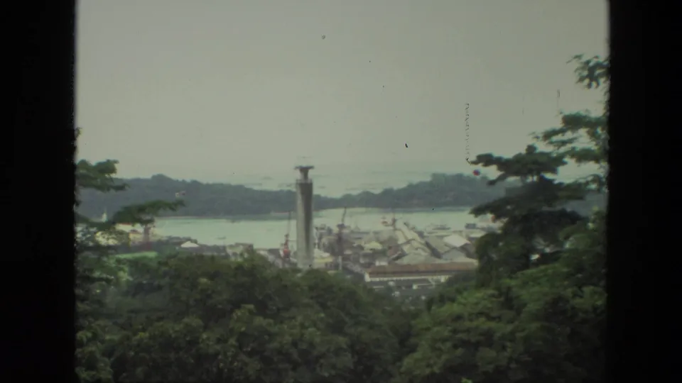 1974 India 8mm frame showing a distant harbor and city structures beyond hillside trees, with a tall tower near the waterfront.